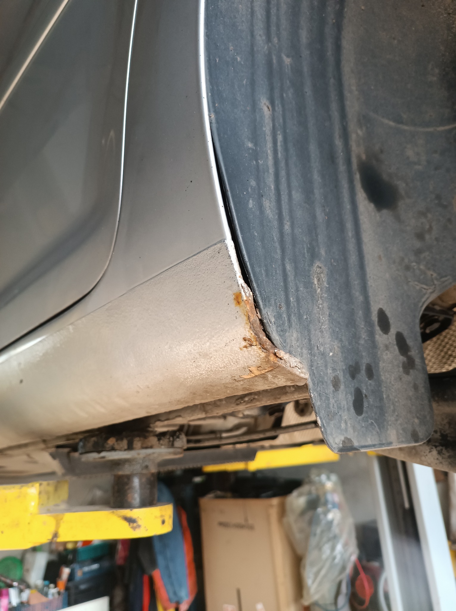 Widok z bliska skorodowanego progu srebrnego samochodu na podnośniku warsztatowym, z widocznymi śladami rdzy i brudu.