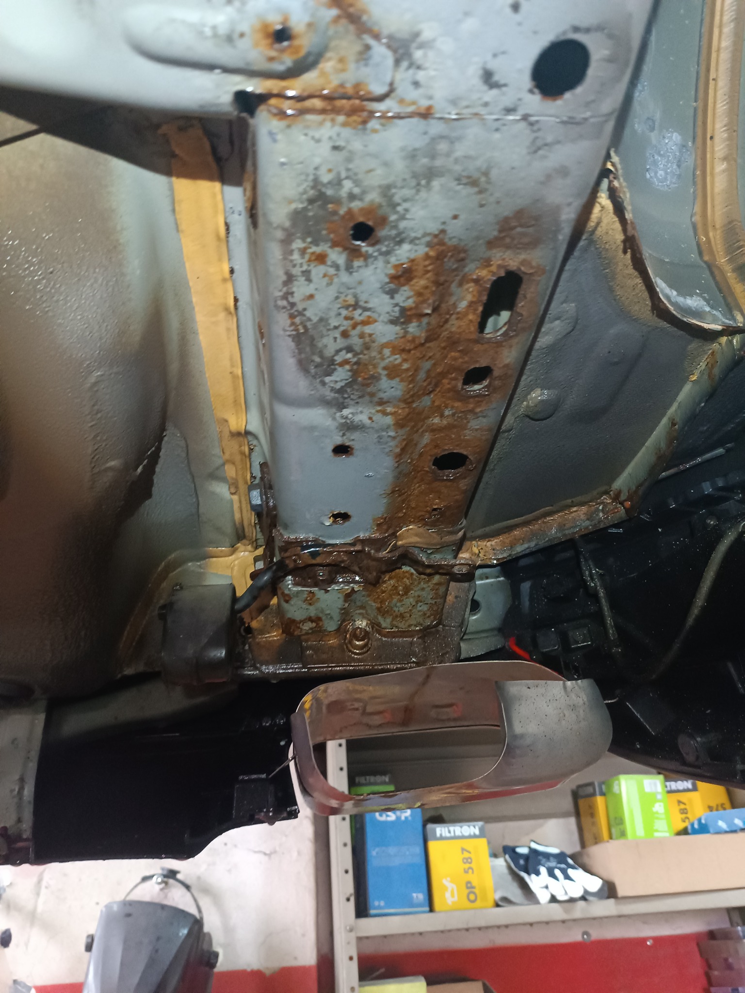 Podwozie samochodu z widocznymi śladami rdzy i korozji, sfotografowane z dołu, z widocznymi elementami konstrukcyjnymi i warsztatowym otoczeniem.