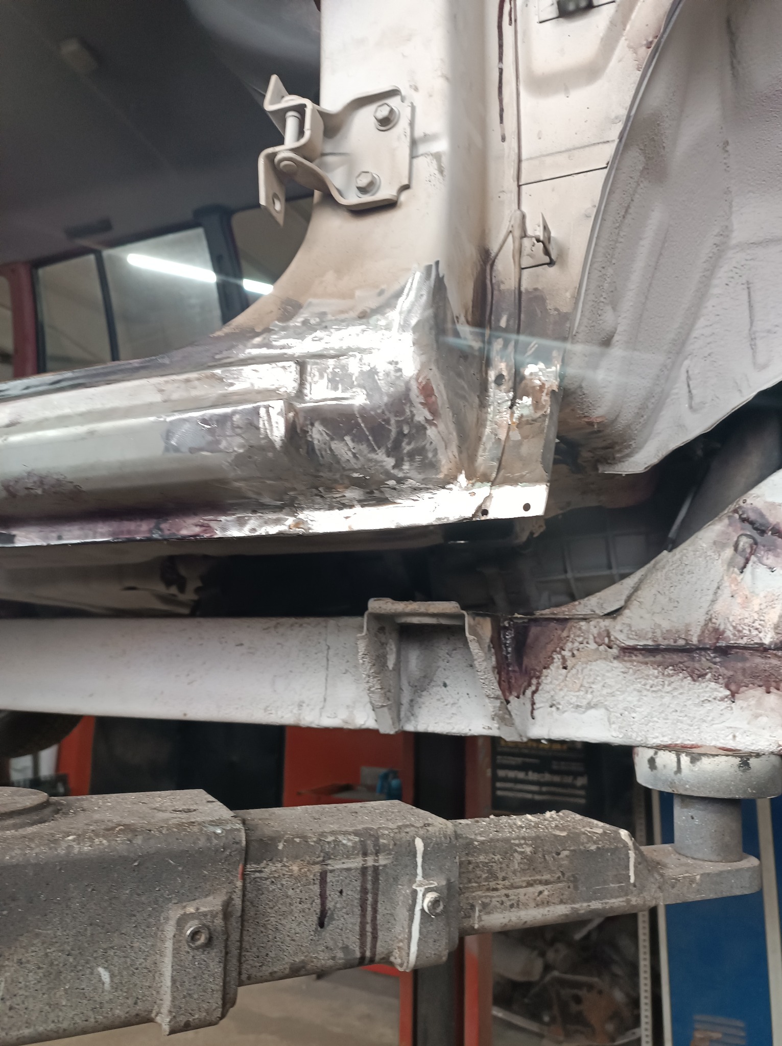 Widok z dołu na fragment karoserii samochodu na podnośniku warsztatowym, z widocznymi śladami napraw blacharskich i zabezpieczenia antykorozyjnego.