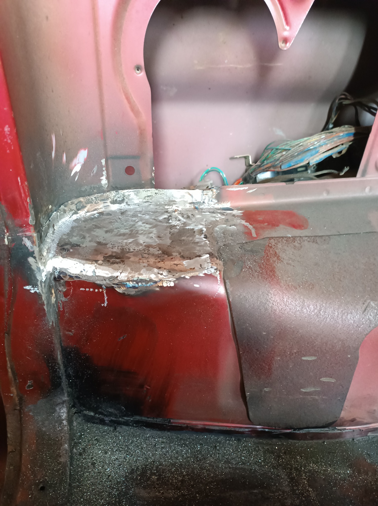 Fragment karoserii samochodu w kolorze czerwonym, z widocznymi śladami napraw blacharskich i spawania, odkryta wiązka przewodów w tle.