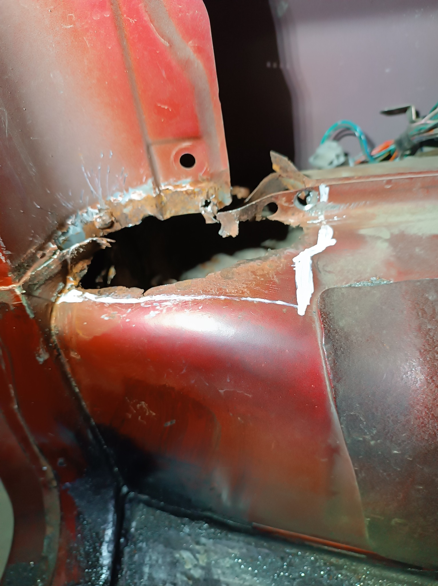 Czerwony element karoserii samochodu z dziurą spowodowaną korozją, w trakcie naprawy z widocznymi śladami rdzy i białym uszczelniaczem.