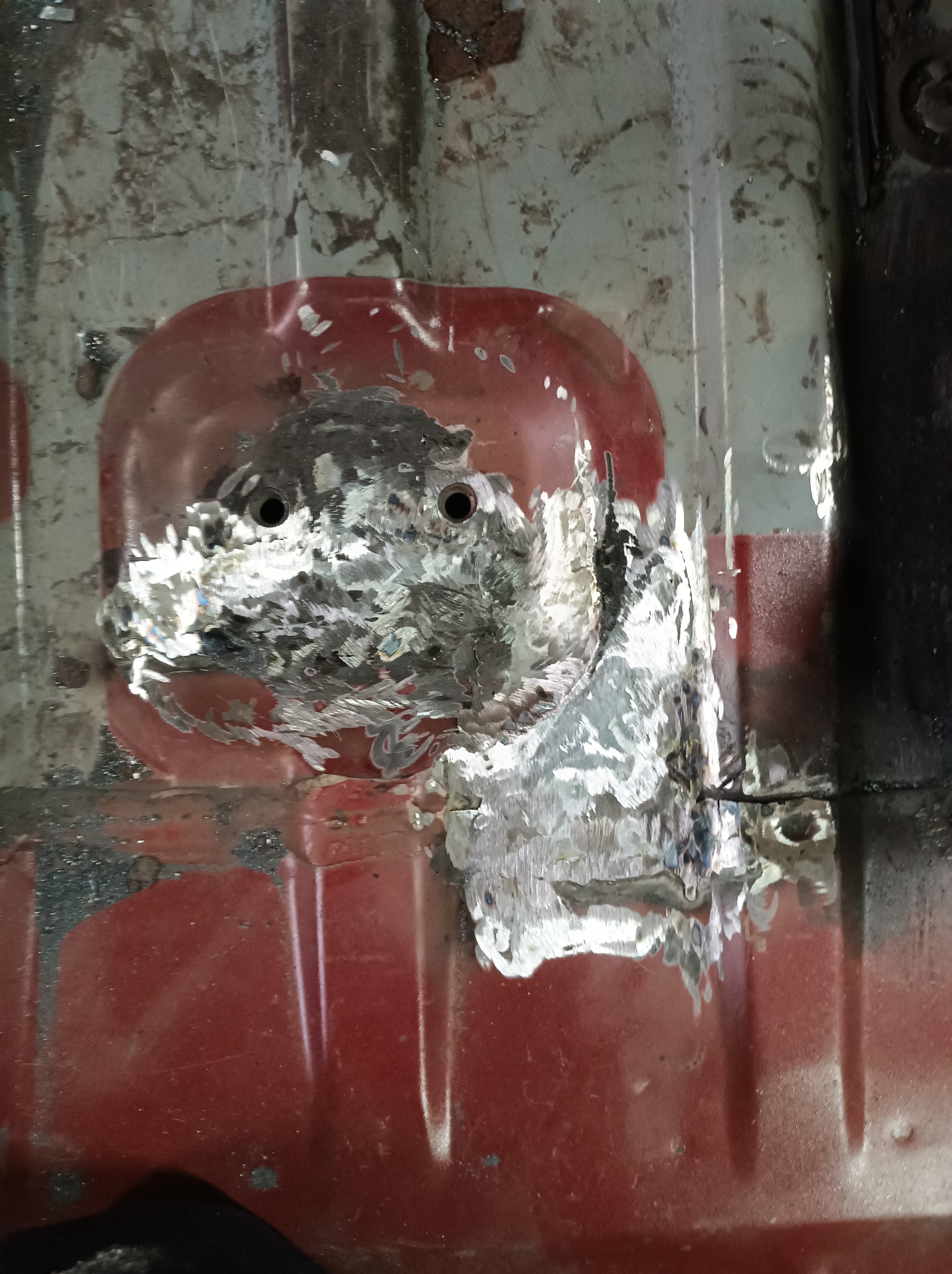 Fragment podwozia samochodu z widocznymi śladami napraw blacharskich i spawania, zarysowania i odpryski farby na czerwonej powierzchni.