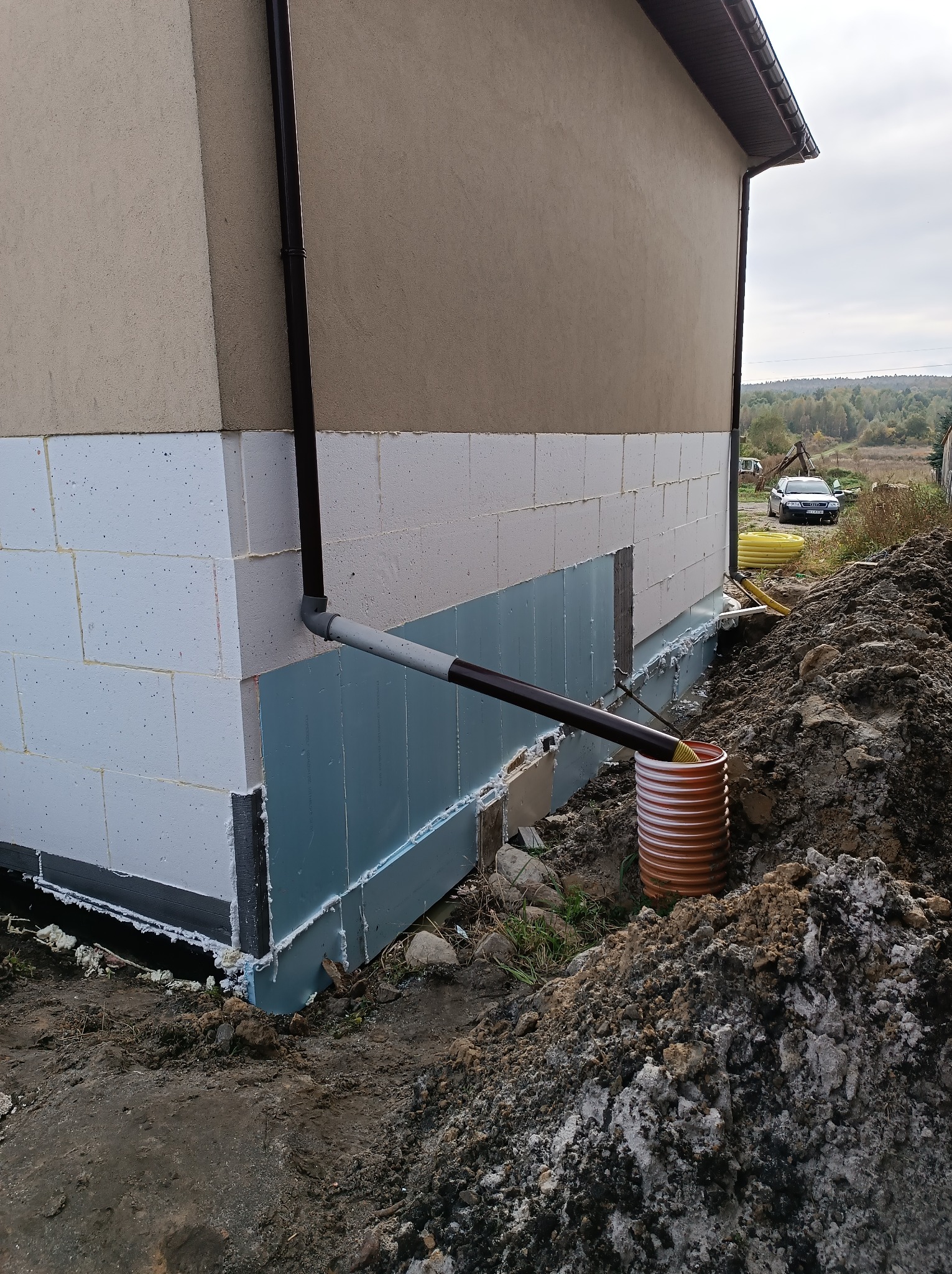 Fragment budynku z widocznym systemem odprowadzania wody deszczowej do pomarańczowej rury drenarskiej w ziemi, ocieplenie fundamentów płytami styropianowymi, tło z krajobrazem i samochodem