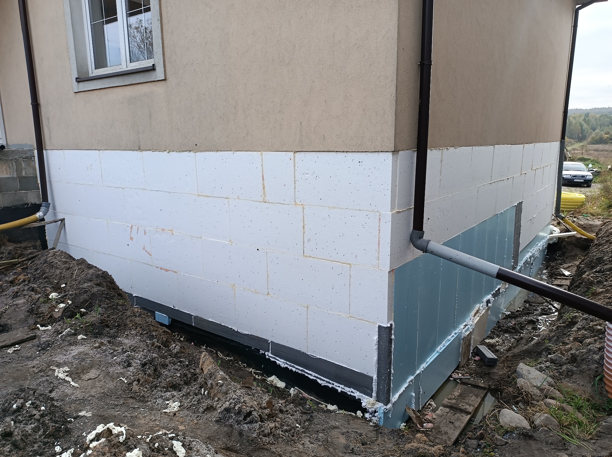 Ocieplenie fundamentów budynku z widoczną izolacją termiczną w postaci białych bloków styropianowych i niebieskich płyt, zabezpieczonych czarną folią hydroizolacyjną, w wykopie ziemnym.