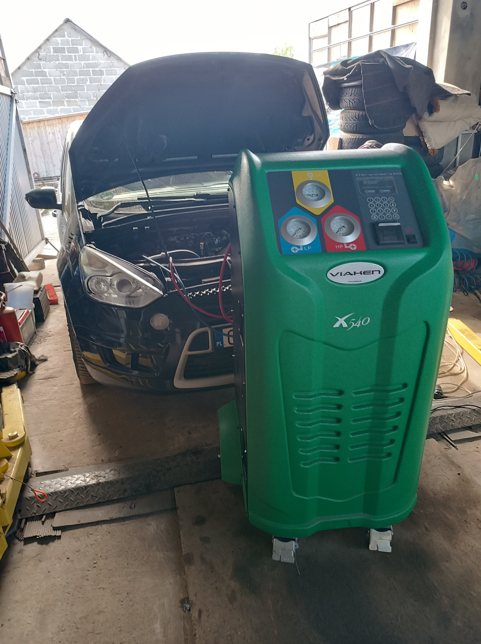 Czarny samochód z otwartą maską na podnośniku warsztatowym, obok stoi zielone urządzenie do obsługi klimatyzacji samochodowej VIAKEN X540 z wyświetlaczem i manometrami.