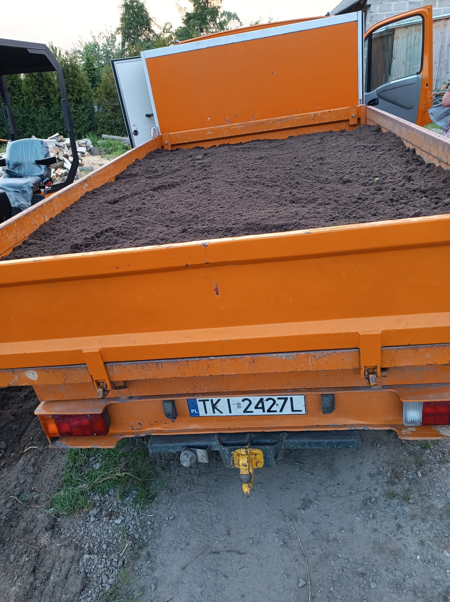 Pomarańczowa wywrotka wypełniona ziemią, widoczny hak holowniczy i tablica rejestracyjna, w tle fragmenty roślinności i pojazd