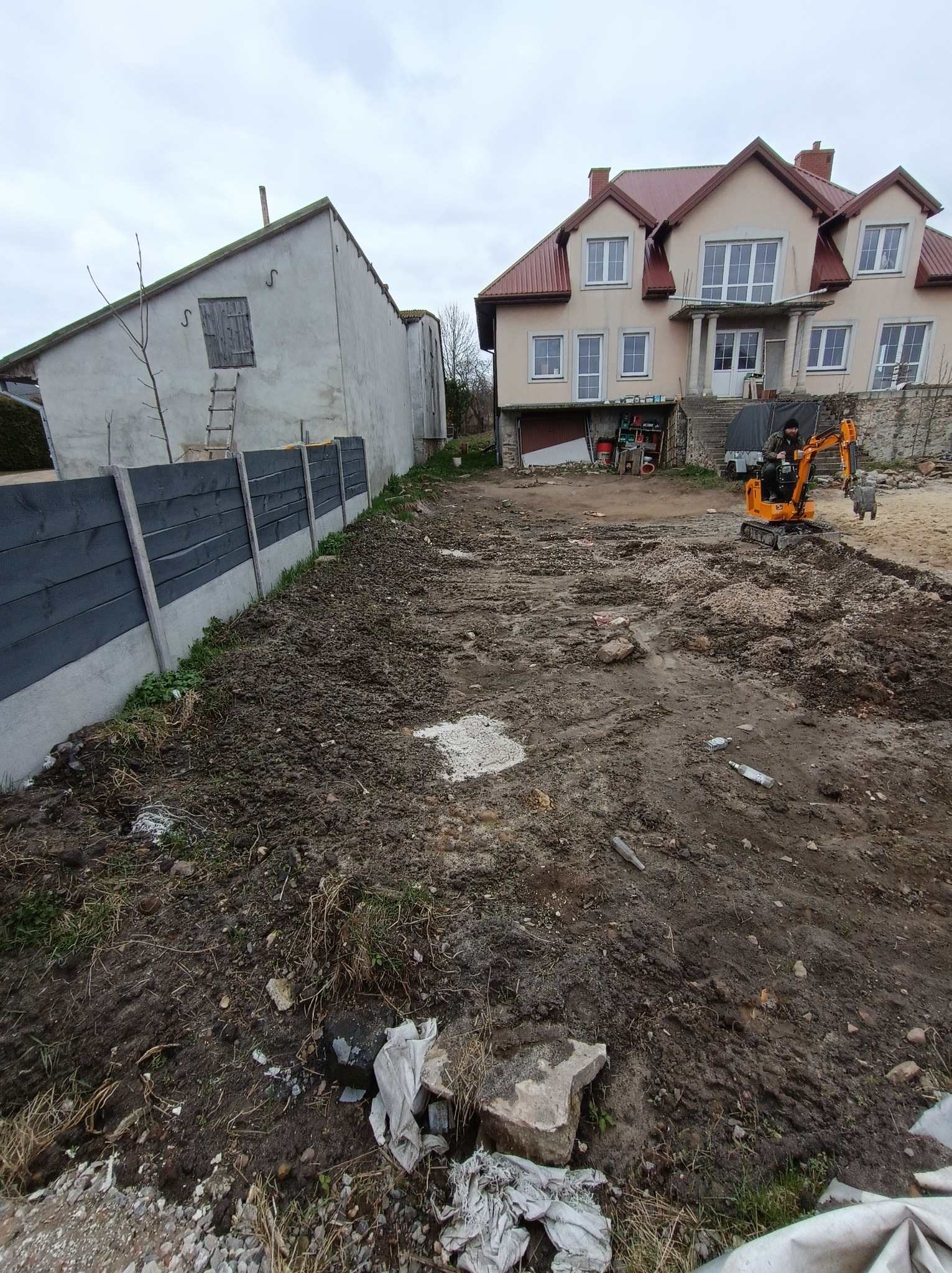 Pomarańczowa mini-koparka na nierównym, przygotowanym terenie budowy z dwoma budynkami w tle, widoczne ślady opon i resztki materiałów budowlanych.