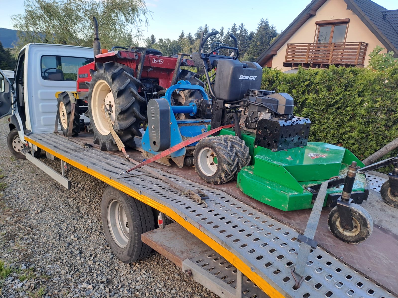 Transport dwóch maszyn ogrodniczych - czerwonego traktora Yanmar i zielonej kosiarki Bob-Cat - na platformie samochodu dostawczego, z domem jednorodzinnym i zielenią w tle.