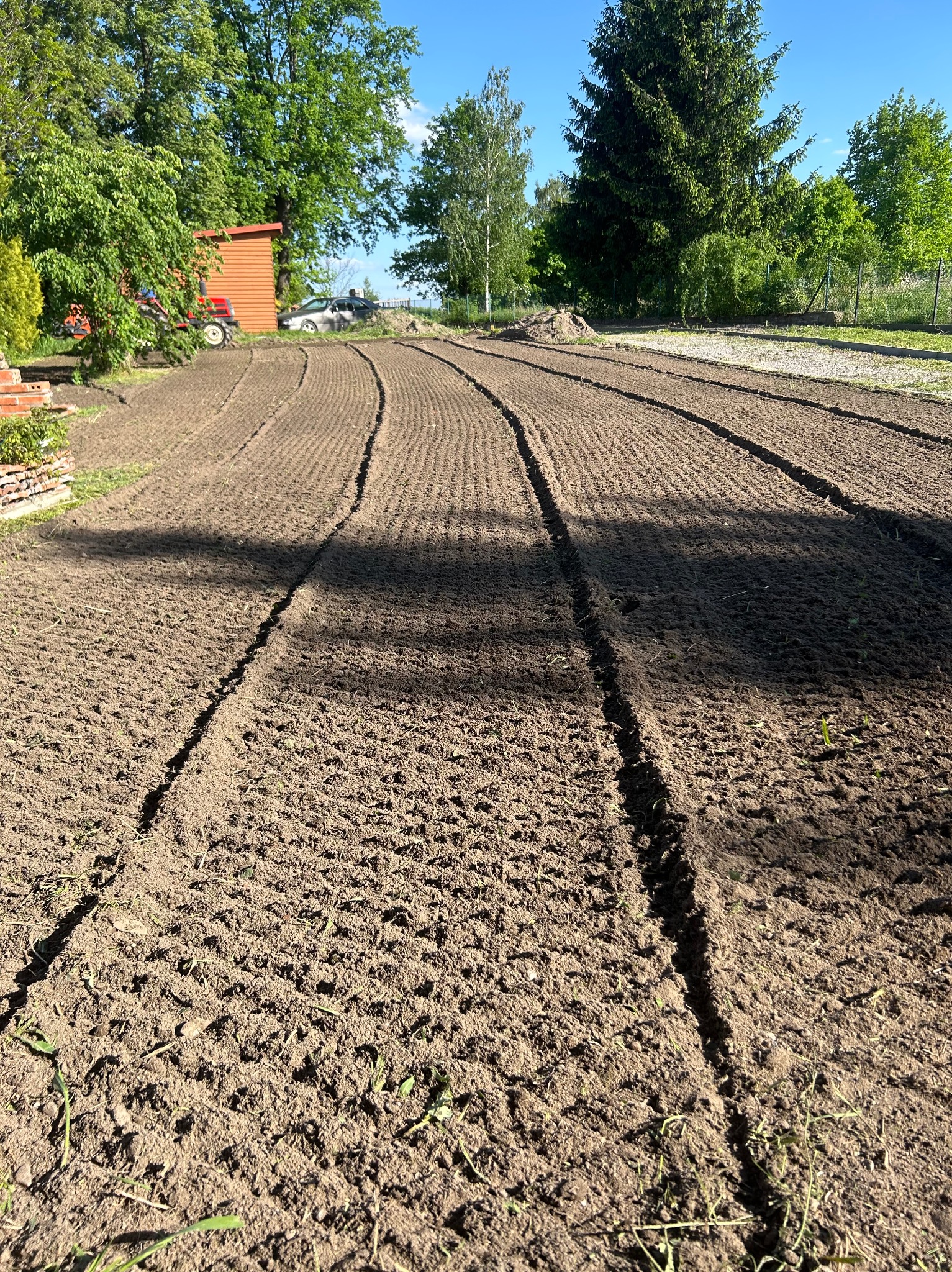 Przygotowana ziemia pod sadzenie z widocznymi bruzdami, w tle budynek gospodarczy i traktor.