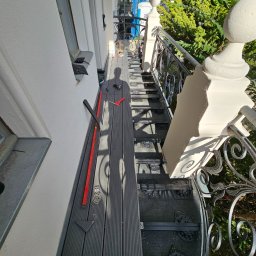 Samuel Talarczyk - Montaż kompozytowej deski tarasowej na schodach zewnętrznych z ozdobną metalową balustradą, widoczne narzędzia i cień osoby montującej.