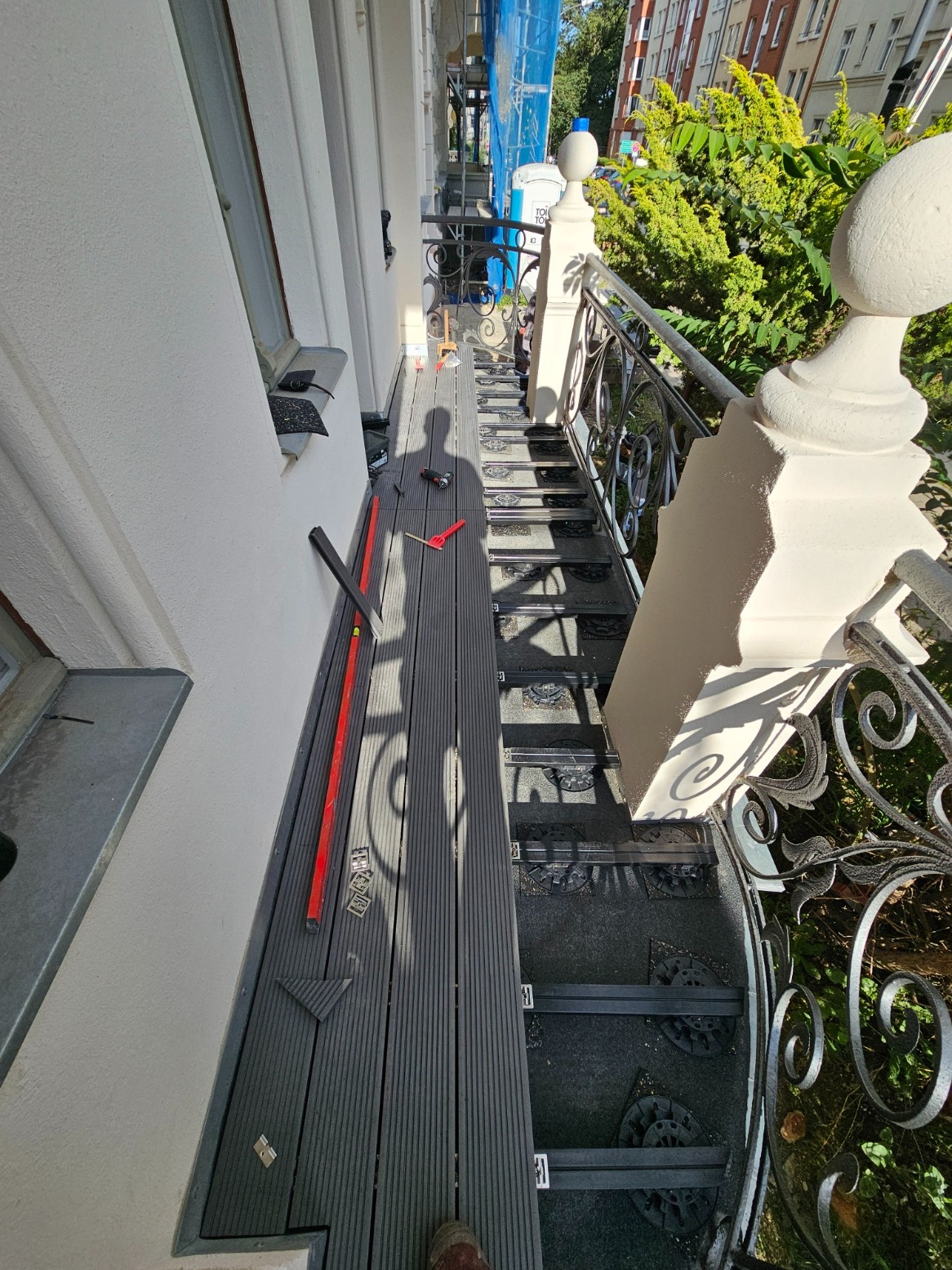 Montaż kompozytowej deski tarasowej na schodach zewnętrznych z ozdobną metalową balustradą, widoczne narzędzia i cień osoby montującej.