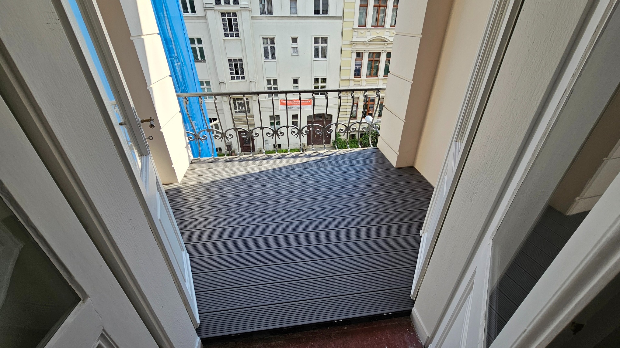 Nowa, szara deska kompozytowa na balkonie z metalową balustradą i widokiem na budynki mieszkalne, uchwycona z wnętrza przez otwarte, białe drzwi balkonowe.