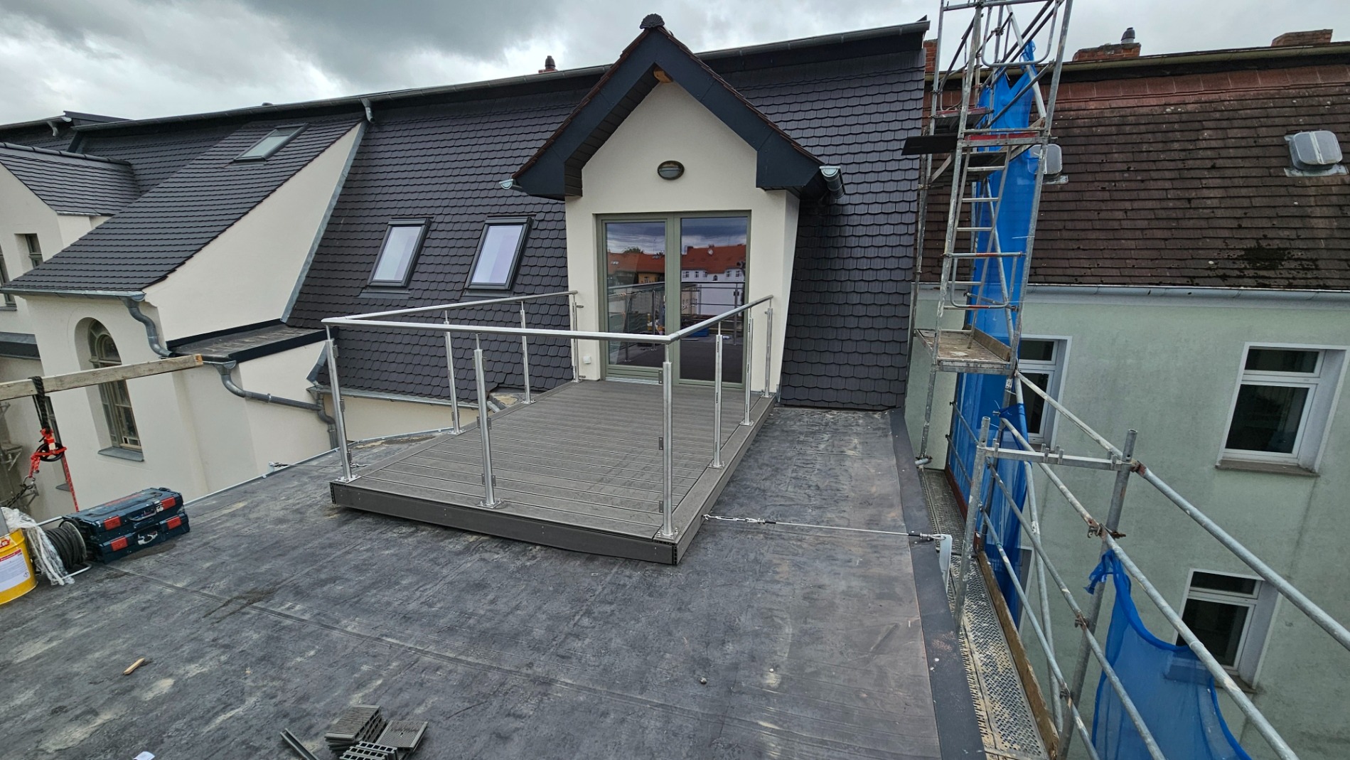 Wykończony dach z balkonem i balustradą ze stali nierdzewnej, widoczne narzędzia i rusztowanie w tle, na dachu czarna papa.