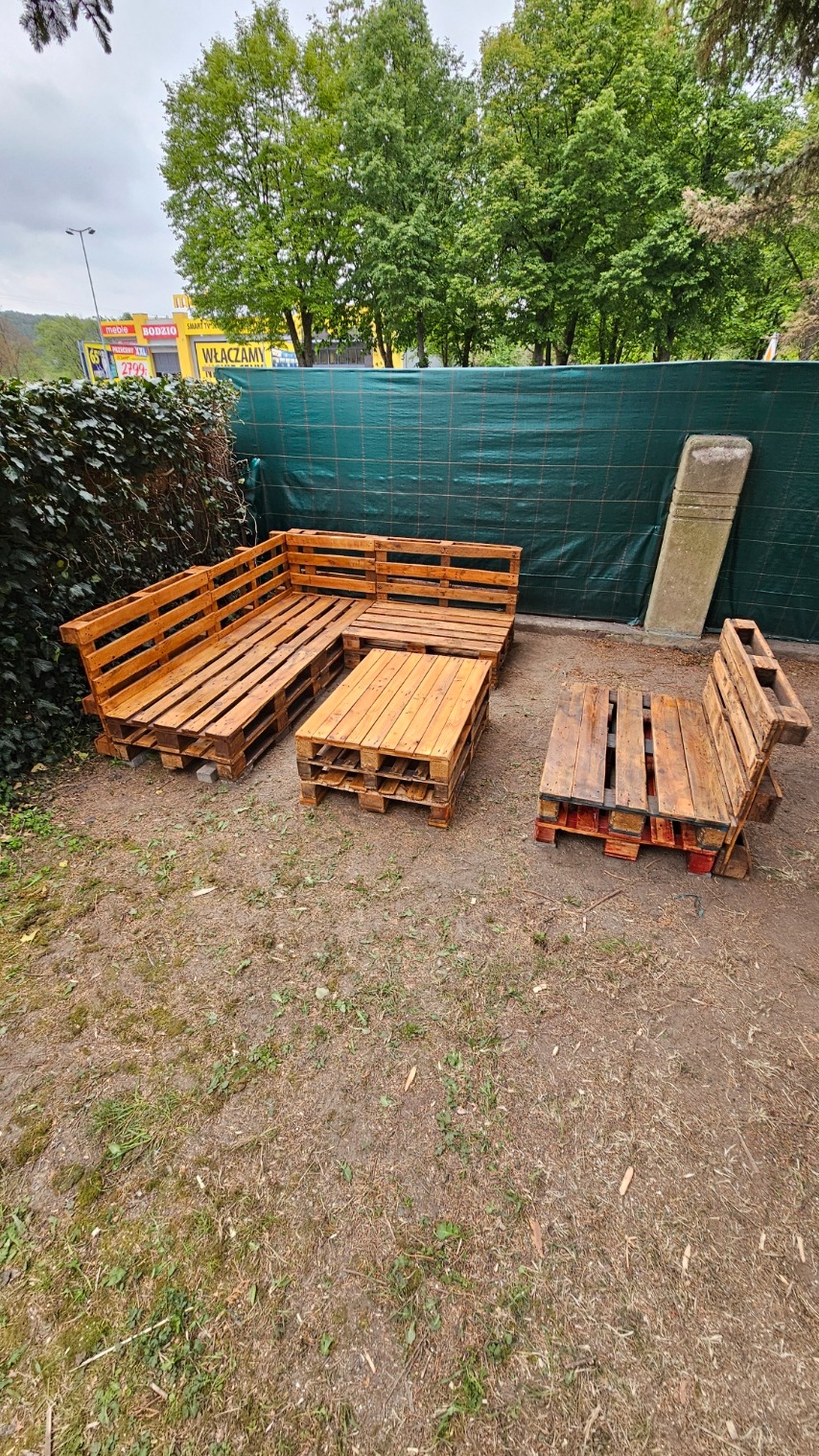 Zestaw mebli ogrodowych z palet: sofa narożna, stolik kawowy i fotel, postawione na trawniku przed zielonym ogrodzeniem.