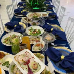 Długi stół bankietowy nakryty granatowym obrusem, ozdobne talerze ze złotymi brzegami, ciemnoniebieskie serwetki ze złotymi pierścieniami, różnorodne dania, napoje w dzbankach i szklankach, białe...