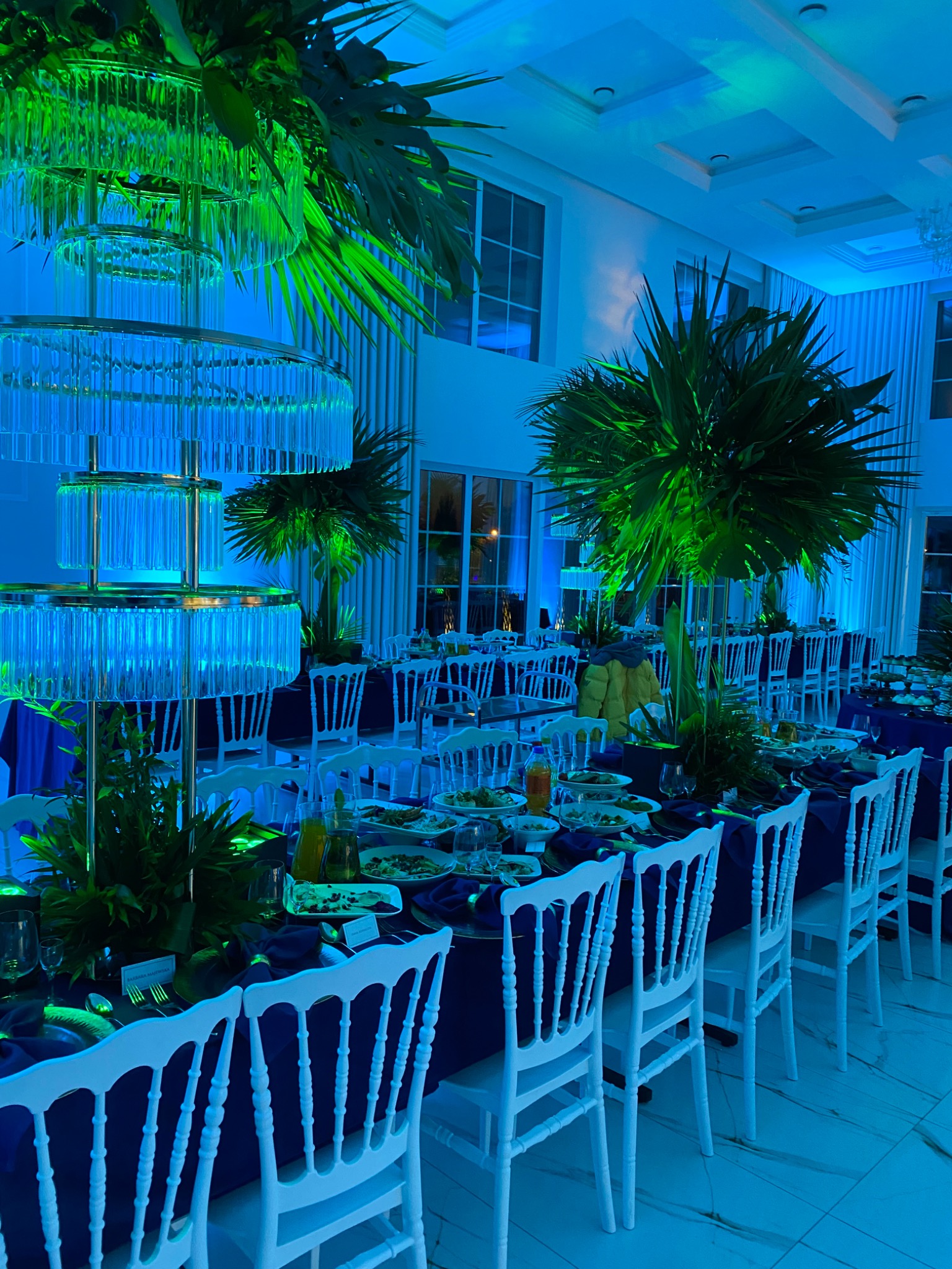 Elegancko nakryty stół na przyjęciu z ciemnoniebieskim obrusem, białymi krzesłami, szklanymi dekoracjami, egzotycznymi roślinami i nowoczesnym oświetleniem w odcieniach zieleni i błękitu.