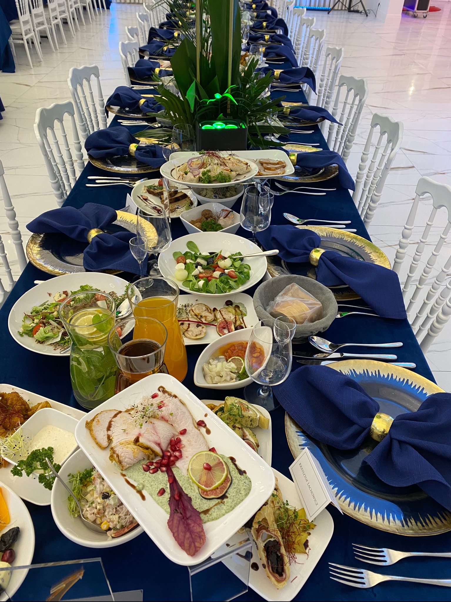 Długi stół bankietowy nakryty granatowym obrusem, ozdobne talerze ze złotymi brzegami, ciemnoniebieskie serwetki ze złotymi pierścieniami, różnorodne dania, napoje w dzbankach i szklankach, białe...