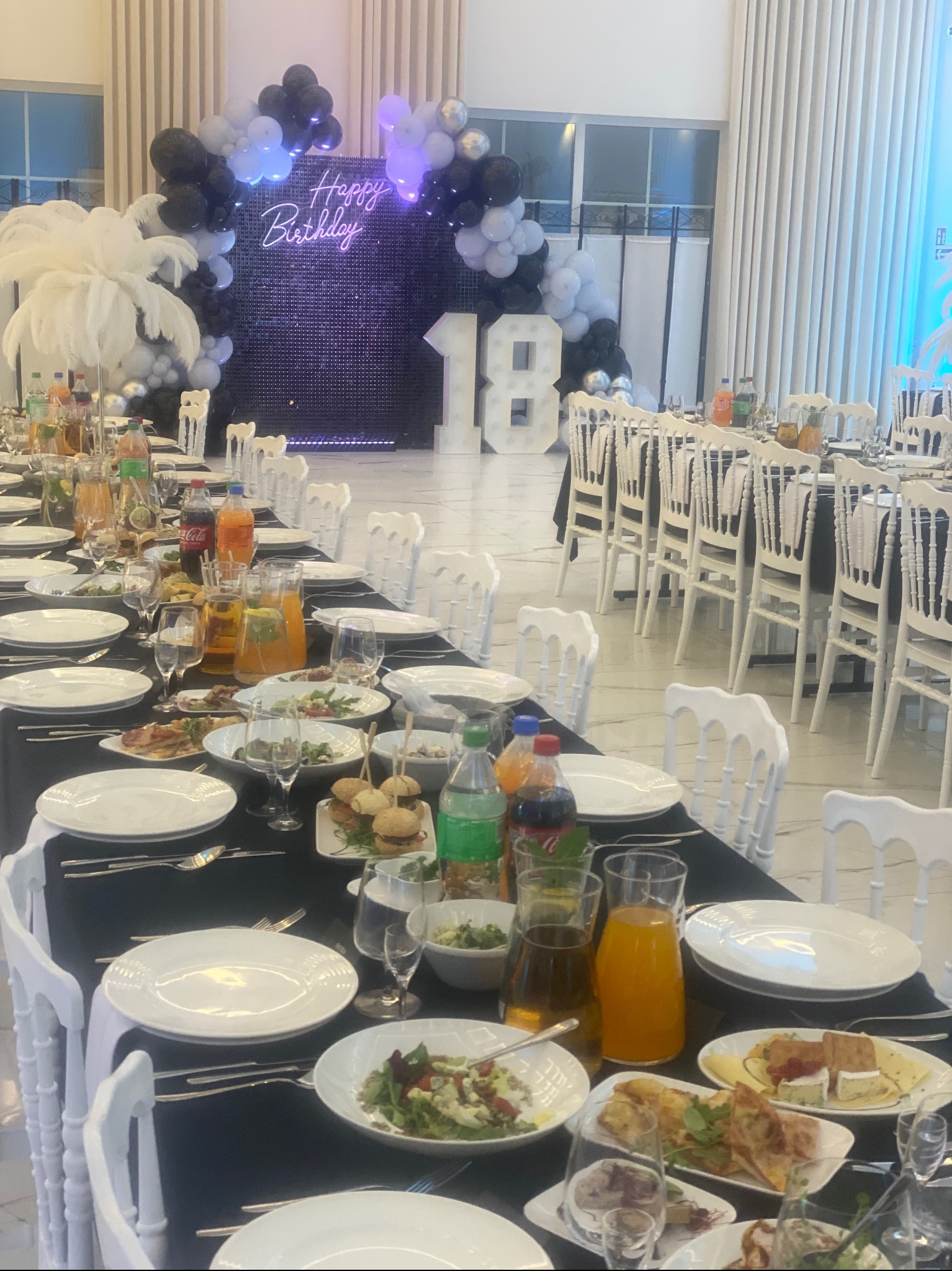 Uroczysta dekoracja sali na przyjęcie urodzinowe: długi stół z czarnym obrusem, nakryty białymi talerzami i sztućcami, z przekąskami, sałatkami i napojami, w tle podświetlony napis 'Happy Birthday'...