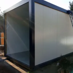 Modułowy kontener mieszkalny w trakcie montażu, widoczna drabina oparta o ścianę i folia zabezpieczająca wewnątrz, w tle drzewa.