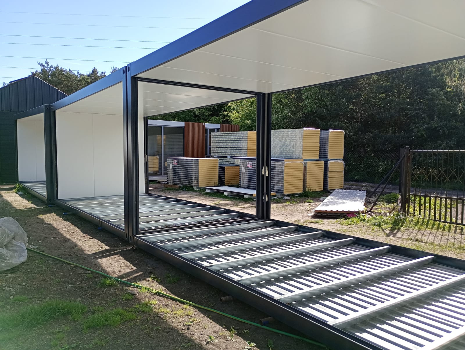 Wstępny etap budowy modułowego domu z widoczną stalową konstrukcją podłogi, ścianami z białych paneli i stosem płyt warstwowych w tle.