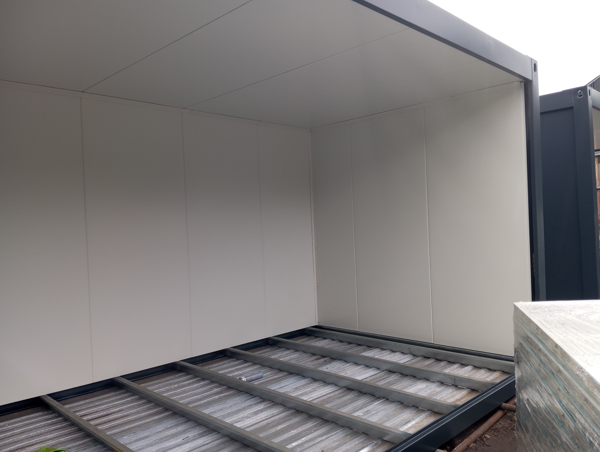 Wnętrze modułu mieszkalnego w trakcie montażu, widoczne ściany z płyt warstwowych i stalowa konstrukcja podłogi, z zewnątrz obudowa z ciemnego metalu.