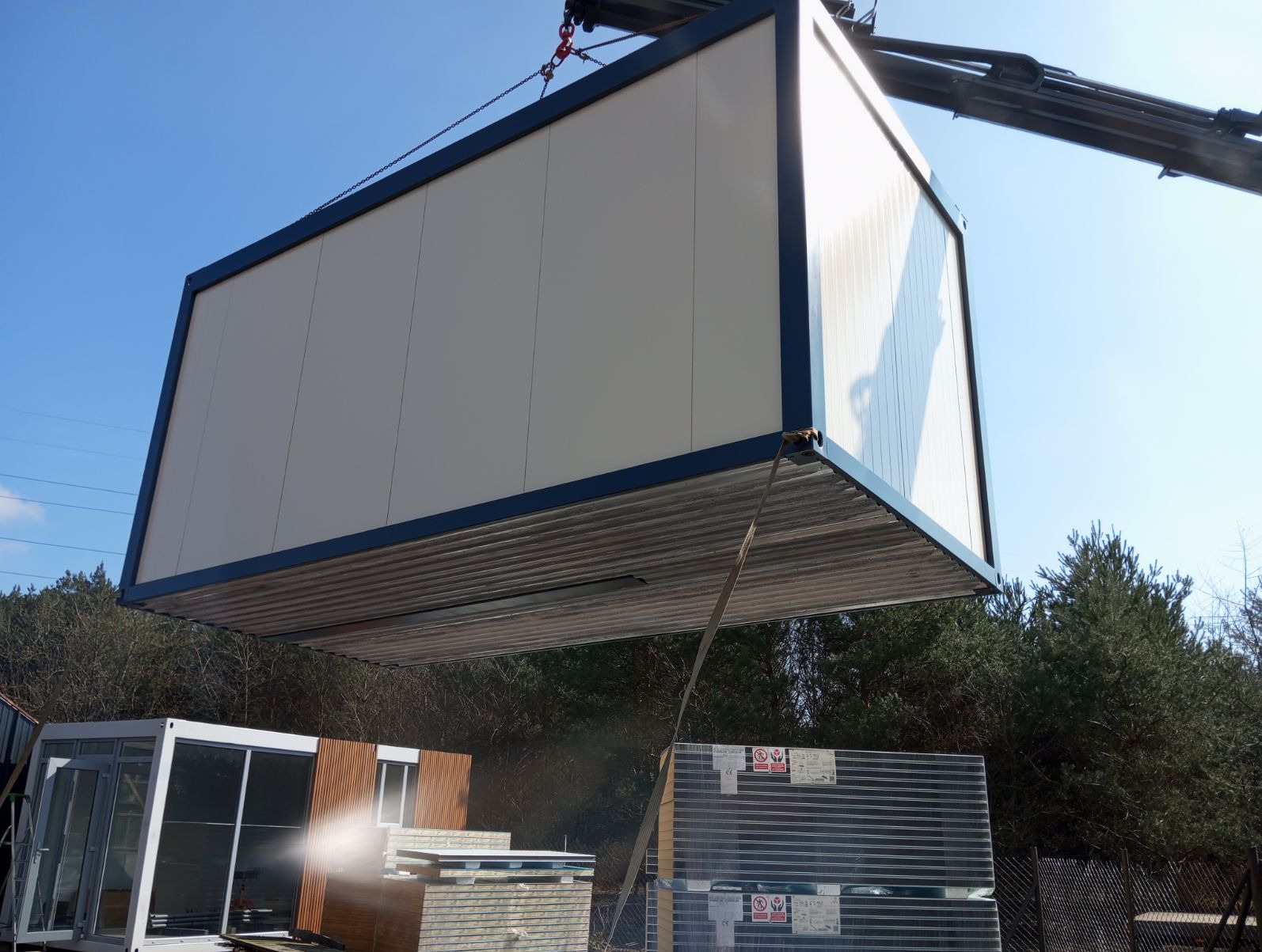 Montaż modułowej konstrukcji z użyciem dźwigu, widoczne stalowe liny mocujące, w tle drzewa i błękitne niebo.