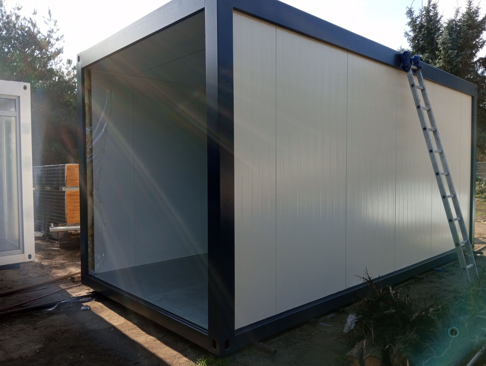 Modułowy kontener mieszkalny w trakcie montażu, widoczna drabina oparta o ścianę i folia zabezpieczająca wewnątrz, w tle drzewa.