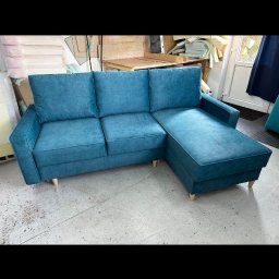 Leńmeble - Turkusowy narożnik z welurową tapicerką i jasnymi drewnianymi nóżkami stoi w warsztacie tapicerskim na betonowej podłodze. W tle widoczne materiały i narzędzia.