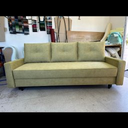 Leńmeble - Prosty, trzyosobowy tapicerowany kanapa w odcieniach zieleni, z trzema poduszkami oparciowymi z guzikami, stoi na betonowej podłodze w warsztacie, w tle próbki materiałów i narzędzia.