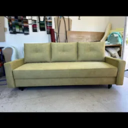 Prosty, trzyosobowy tapicerowany kanapa w odcieniach zieleni, z trzema poduszkami oparciowymi z guzikami, stoi na betonowej podłodze w warsztacie, w tle próbki materiałów i narzędzia.