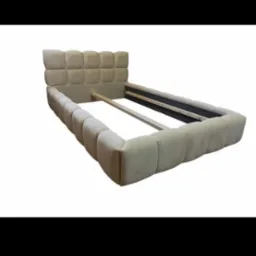 Rama tapicerowanego łóżka w kolorze beżowym z pikowanym zagłówkiem, widoczne drewniane stelaże, białe tło.