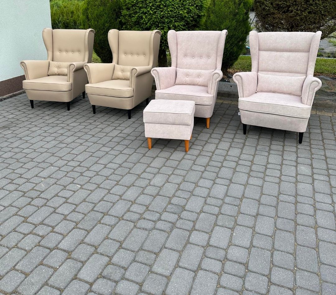 Dwa beżowe i dwa różowe fotele tapicerowane typu uszak z podnóżkiem, ustawione na szarej kostce brukowej, z zielonymi krzewami w tle. Fotele posiadają charakterystyczne wysokie oparcia i poduszki.