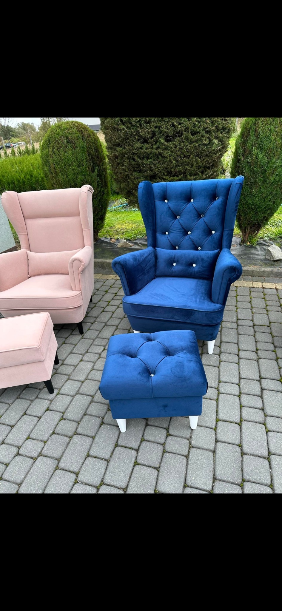 Komplet wypoczynkowy: fotel typu uszak w kolorze pudrowego różu oraz fotel uszak i pufa w kolorze granatowym, oba wykończone guzikami, na tle brukowanej nawierzchni i zieleni.