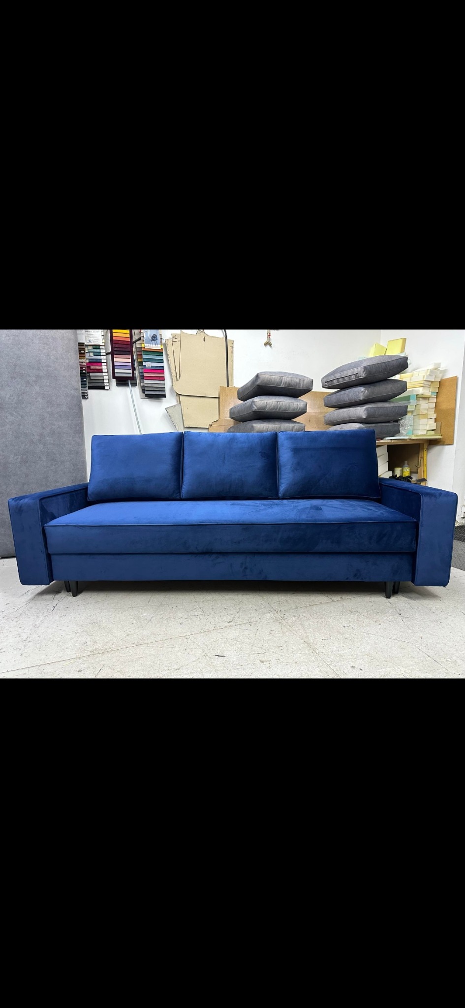 Nowoczesna, granatowa sofa z trzema poduszkami na tle warsztatu tapicerskiego z próbkami tkanin i stosem poduszek.