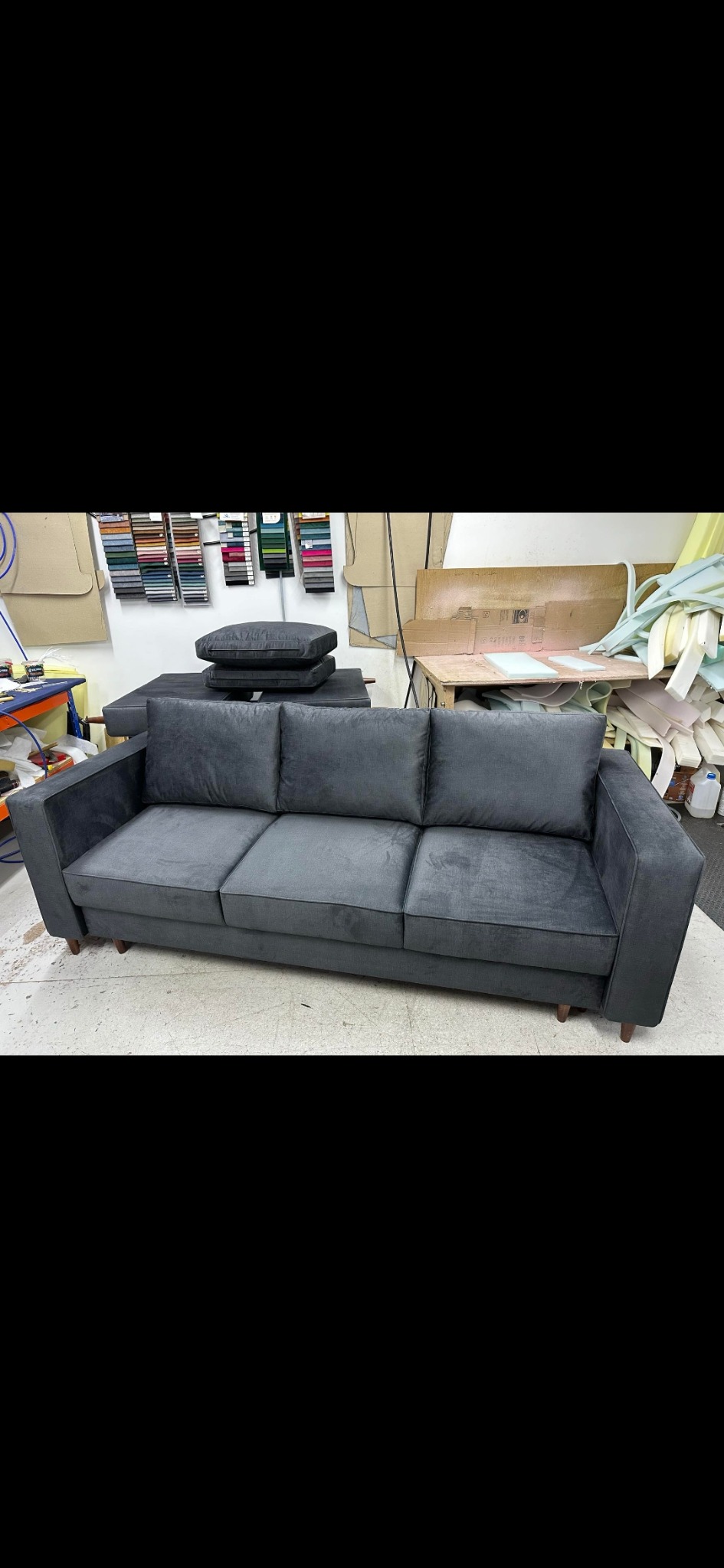 Grafitowa sofa trzyosobowa z poduszkami na tle warsztatu tapicerskiego z próbkami tkanin i narzędziami.