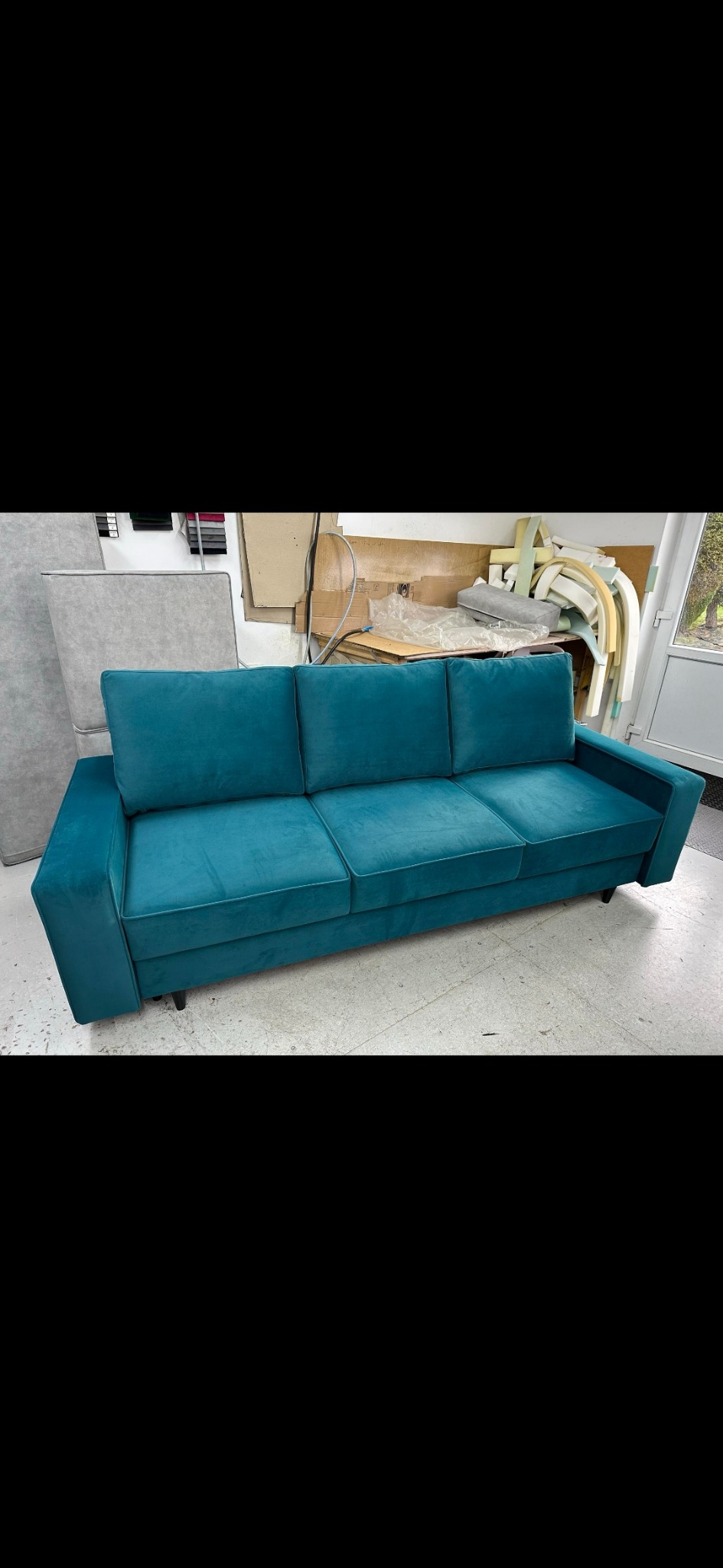 Turkusowa sofa trzyosobowa z weluru w warsztacie tapicerskim, widoczne materiały i narzędzia w tle.