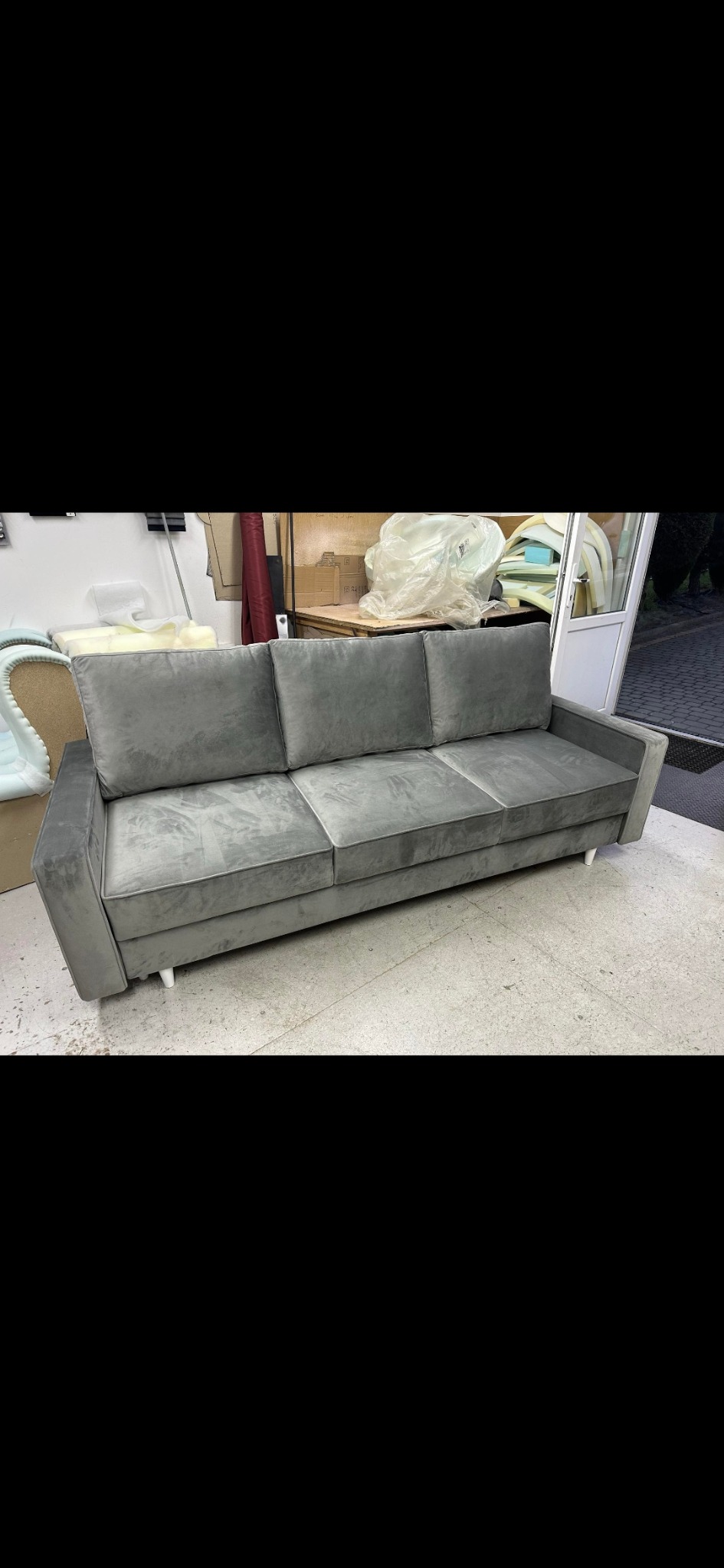 Nowoczesna, szara sofa trzyosobowa z welurową tapicerką i minimalistycznymi nóżkami, sfotografowana w warsztacie tapicerskim z widocznymi materiałami i narzędziami w tle.