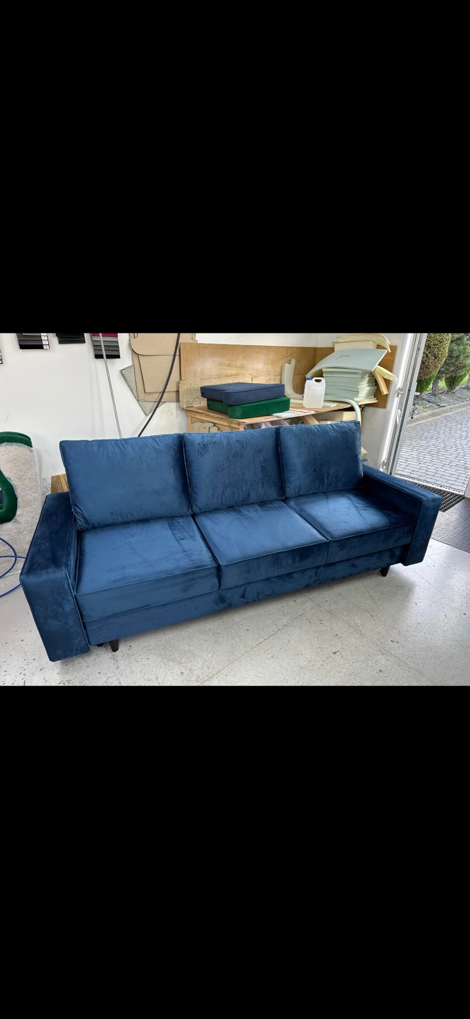 Granatowa, welurowa sofa trzyosobowa na niskich, czarnych nóżkach, sfotografowana w warsztacie tapicerskim z widocznymi materiałami i narzędziami w tle.
