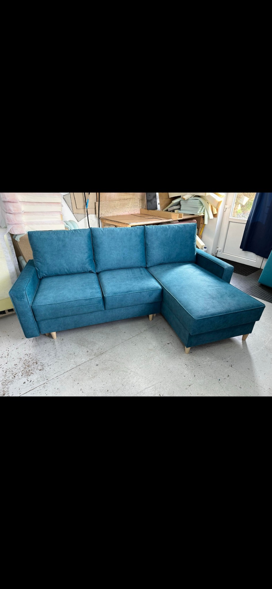 Turkusowy narożnik z welurową tapicerką i jasnymi drewnianymi nóżkami stoi w warsztacie tapicerskim na betonowej podłodze. W tle widoczne materiały i narzędzia.