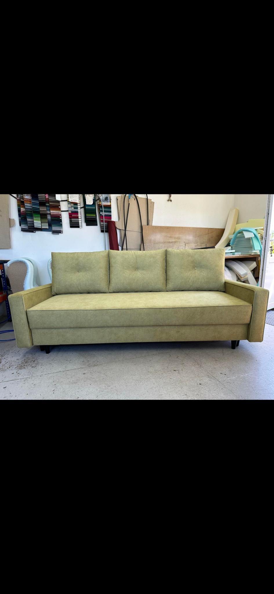 Prosty, trzyosobowy tapicerowany kanapa w odcieniach zieleni, z trzema poduszkami oparciowymi z guzikami, stoi na betonowej podłodze w warsztacie, w tle próbki materiałów i narzędzia.