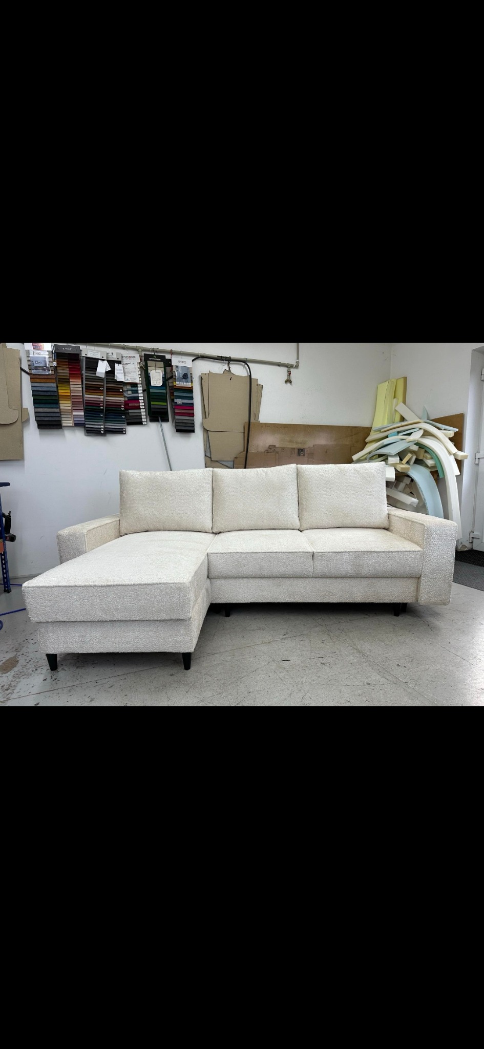 Jasna, narożna kanapa z grubą, teksturowaną tapicerką w warsztacie, widoczne próbki materiałów i elementy piankowe w tle.