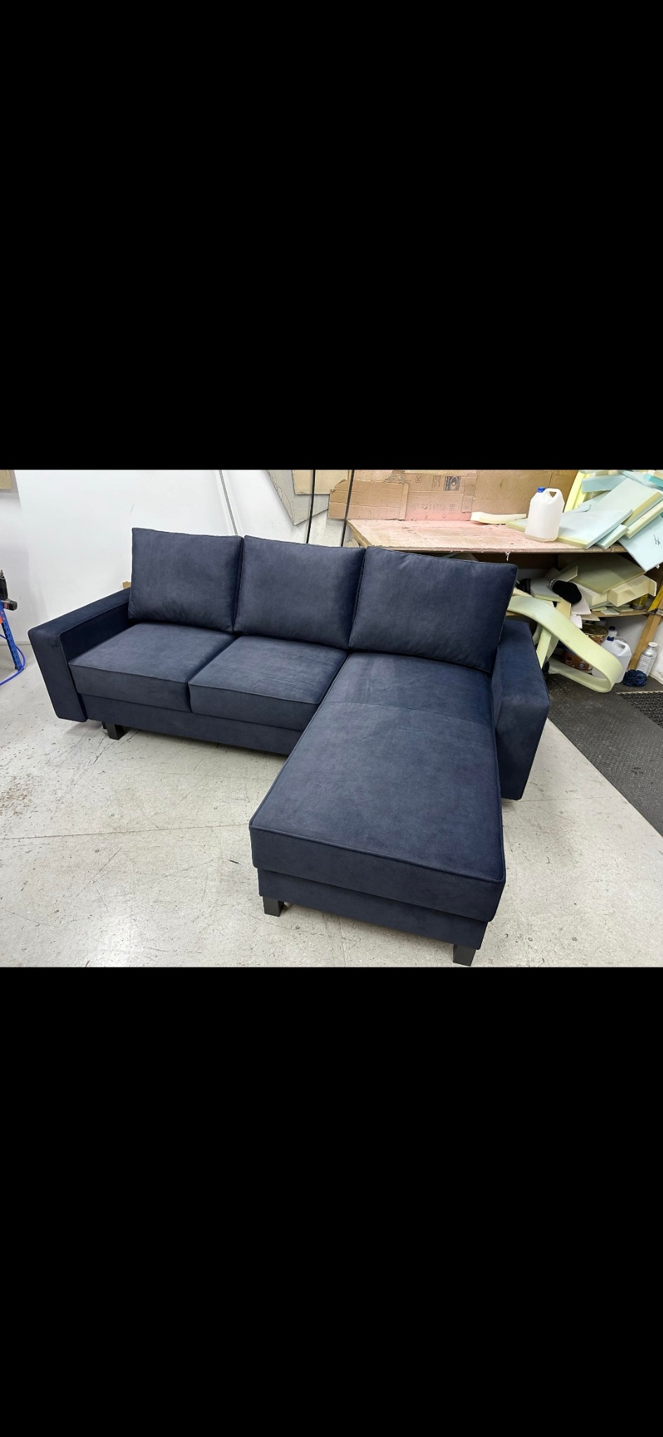 Nowoczesna, narożna sofa w odcieniach granatu z niskimi, kwadratowymi nóżkami, sfotografowana w warsztacie tapicerskim, z widocznymi materiałami i narzędziami w tle.