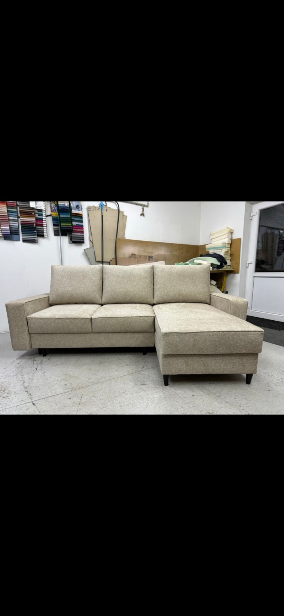 Beżowa sofa narożna z krótkim szezlongiem, ustawiona w warsztacie tapicerskim, widoczne próbki tkanin na ścianie i materiały tapicerskie w tle.