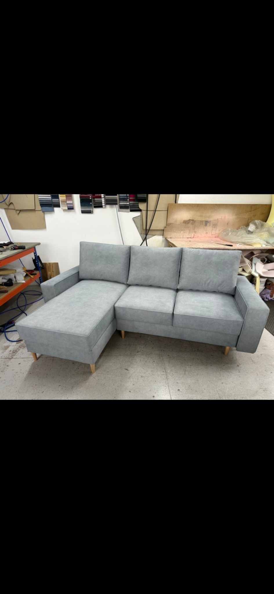 Jasnoszara sofa narożna z drewnianymi nóżkami w warsztacie tapicerskim, widoczne próbki tkanin na ścianie i narzędzia pracy.