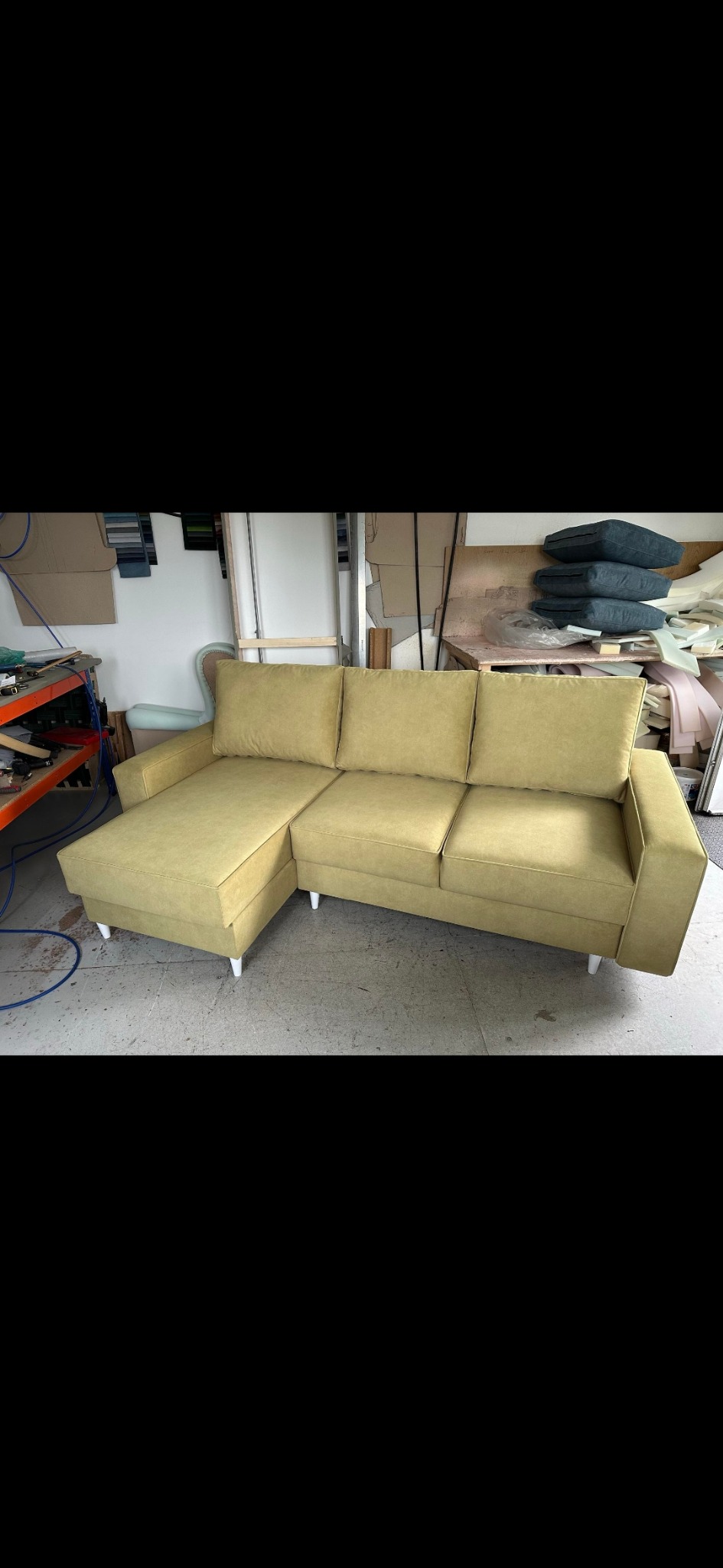 Jasnozielona sofa narożna z trzema poduszkami o prostym kroju i białymi nóżkami, w tle warsztat tapicerski z narzędziami i materiałami.