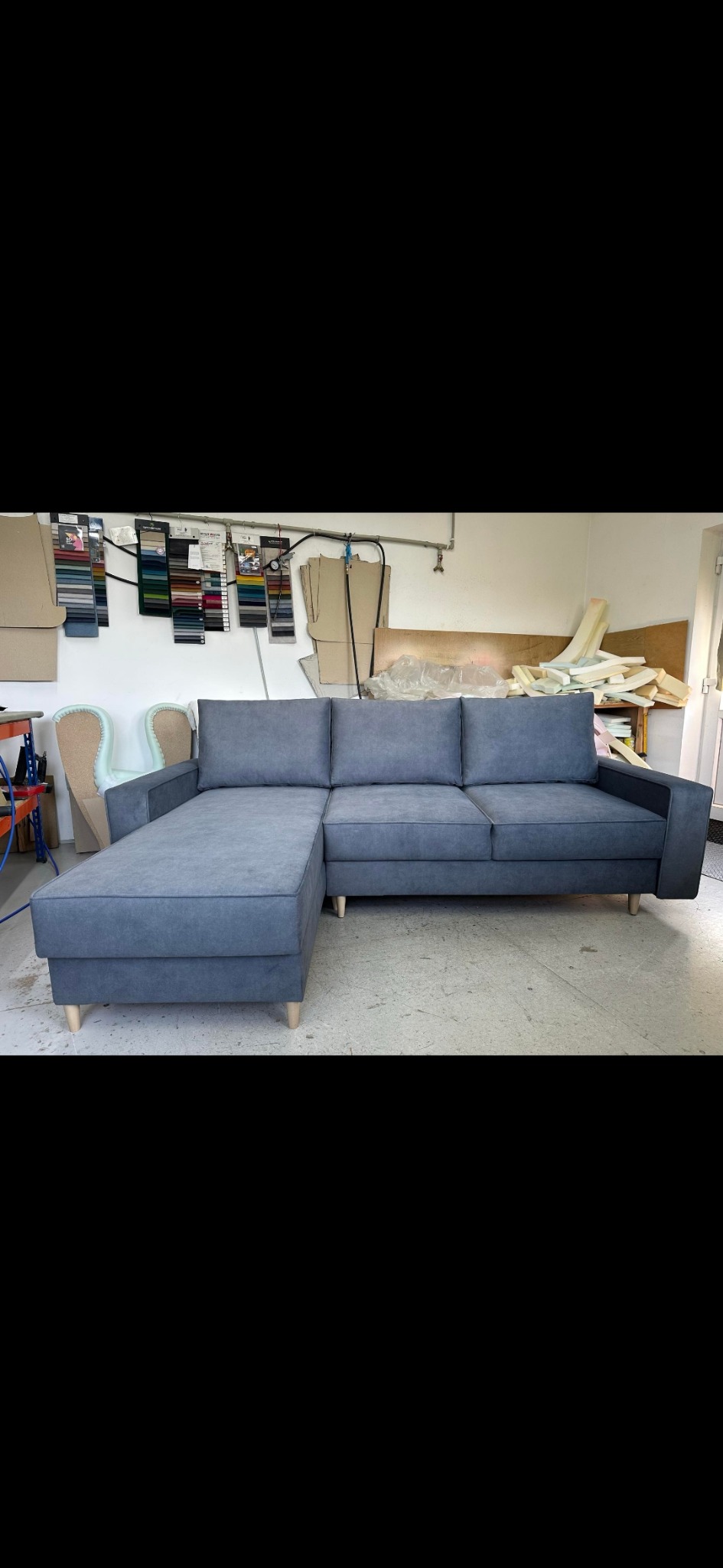 Szara sofa narożna na drewnianych nóżkach w warsztacie tapicerskim, widoczne próbki tkanin i materiały tapicerskie w tle.