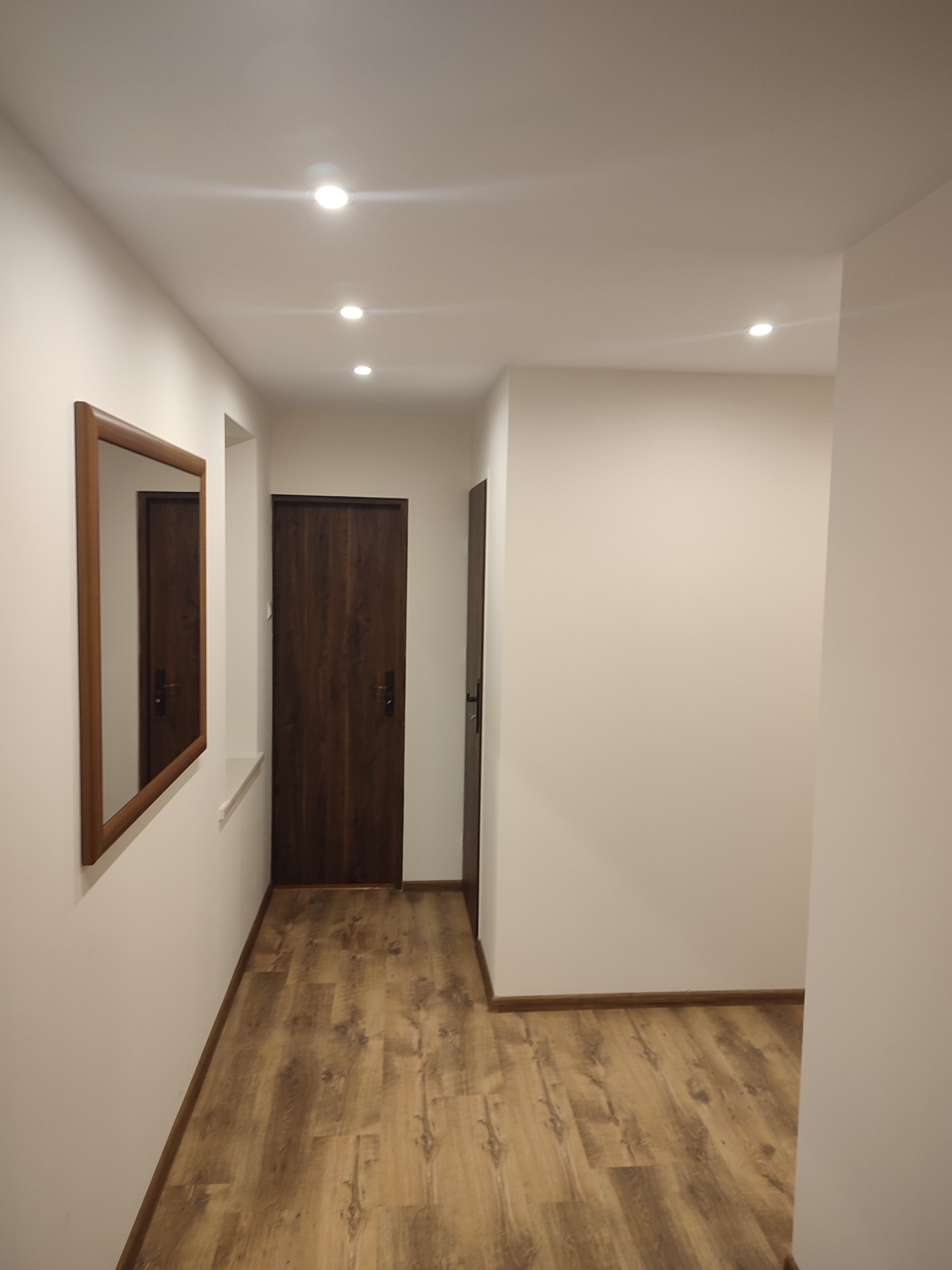 Jasny korytarz po remoncie: drewnopodobne panele, świeżo pomalowane ściany w odcieniach bieli, lustro w drewnianej ramie i dwoje ciemnych drzwi.