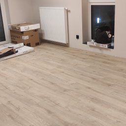 Osoba - Jasne panele podłogowe ułożone w nowym mieszkaniu, widoczne kartony z panelami i grzejnik na ścianie, torba i butelka na parapecie okna.