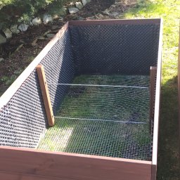Osoba - Drewniana skrzynia na warzywa z wewnętrzną wyściółką z tworzywa sztucznego i siatką zabezpieczającą przed gryzoniami, ustawiona na trawniku w ogrodzie.