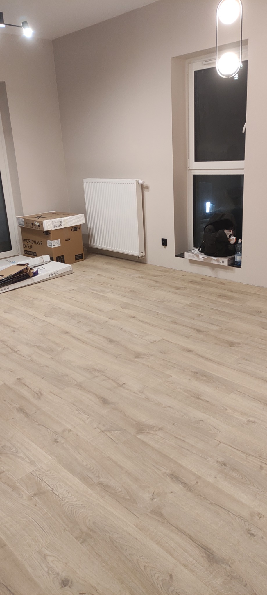 Jasne panele podłogowe ułożone w nowym mieszkaniu, widoczne kartony z panelami i grzejnik na ścianie, torba i butelka na parapecie okna.